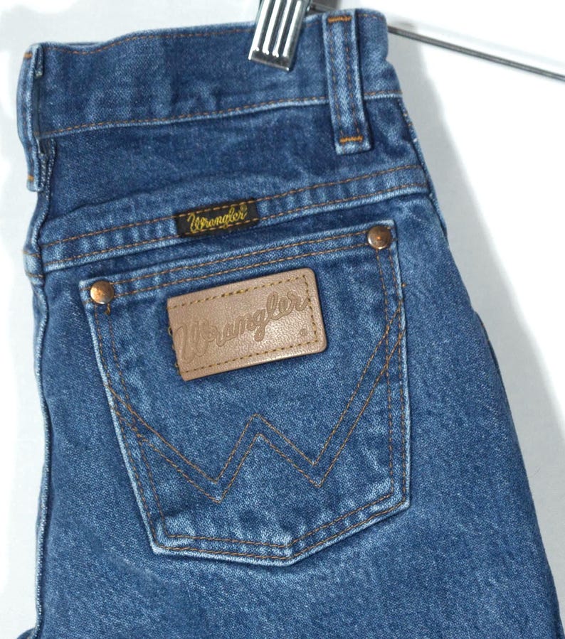 Puede incluir: Primer plano de unos vaqueros de mezclilla azules con un parche de cuero marr&oacute;n en el bolsillo trasero. El parche y la etiqueta peque&ntilde;a llevan el nombre de la marca "Wrangler". Los vaqueros cuelgan de una percha met&aacute;lica.