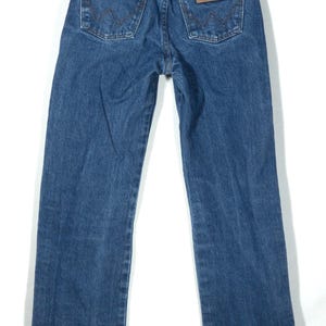 Puede incluir: Vista trasera de unos vaqueros de mezclilla azules con el logo de Wrangler en el bolsillo trasero. Los vaqueros tienen un estilo cl&aacute;sico de cinco bolsillos, un parche de cuero marr&oacute;n y cierre de cremallera y bot&oacute;n met&aacute;licos. Los vaqueros cuelgan sobre un fondo blanco.