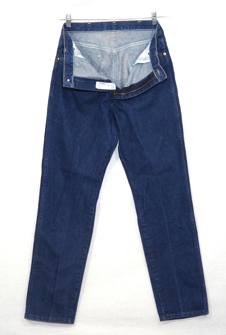 May include: Dark blue denim jeans hanging on a metal hanger. The jeans are unbuttoned and unzipped, revealing the interior lining. The jeans have a classic five-pocket design and a straight leg cut.