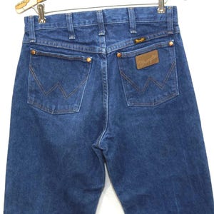 May include: Dark blue denim jeans with the Wrangler logo on the back pocket. The jeans have a classic five-pocket design with copper rivets and a brown leather patch. The back pockets feature the signature Wrangler "W" stitching.