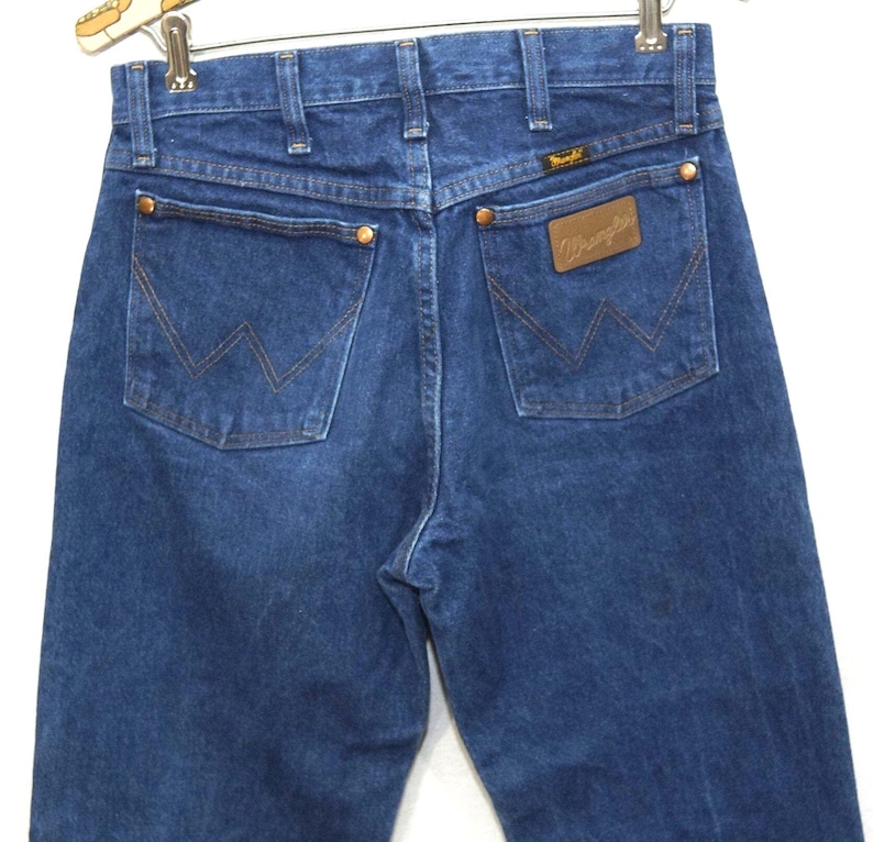 May include: Dark blue denim jeans with the Wrangler logo on the back pocket. The jeans have a classic five-pocket design with copper rivets and a brown leather patch. The back pockets feature the signature Wrangler "W" stitching.
