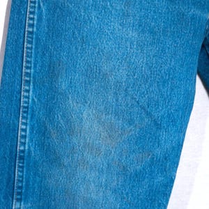 Puede incluir: Primer plano de unos vaqueros de mezclilla azules. Los vaqueros tienen un dise&ntilde;o cl&aacute;sico de cinco bolsillos y un corte recto. La mezclilla tiene un aspecto descolorido con algunas &aacute;reas m&aacute;s oscuras, lo que sugiere desgaste. Las costuras son visibles a lo largo de las costuras.
