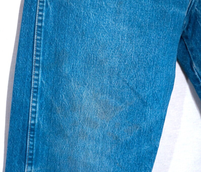 Puede incluir: Primer plano de unos vaqueros de mezclilla azules. Los vaqueros tienen un dise&ntilde;o cl&aacute;sico de cinco bolsillos y un corte recto. La mezclilla tiene un aspecto descolorido con algunas &aacute;reas m&aacute;s oscuras, lo que sugiere desgaste. Las costuras son visibles a lo largo de las costuras.