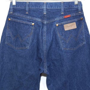 May include: Dark blue denim jeans with the Wrangler logo on the back pocket. The jeans have a classic five-pocket design with brass rivets and a red Wrangler patch. The back pockets feature the signature Wrangler 'W' stitching.