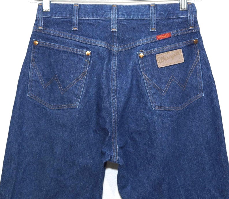 May include: Dark blue denim jeans with the Wrangler logo on the back pocket. The jeans have a classic five-pocket design with brass rivets and a red Wrangler patch. The back pockets feature the signature Wrangler 'W' stitching.