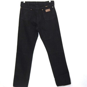 May include: Black denim jeans with a classic five-pocket design. The jeans feature copper rivets, a brown leather patch, and a Wrangler label. The jeans are displayed on a white background.