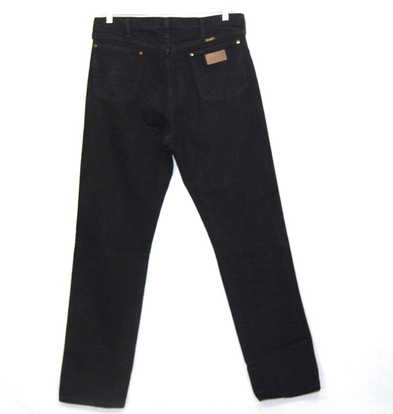 May include: Black denim jeans with a classic five-pocket design. The jeans feature copper rivets, a brown leather patch, and a Wrangler label. The jeans are displayed on a white background.