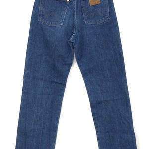 May include: Back view of blue denim jeans with the Wrangler logo on the back pocket and a brown leather patch. The jeans have classic five-pocket styling and are hanging on a vintage patterned hanger.