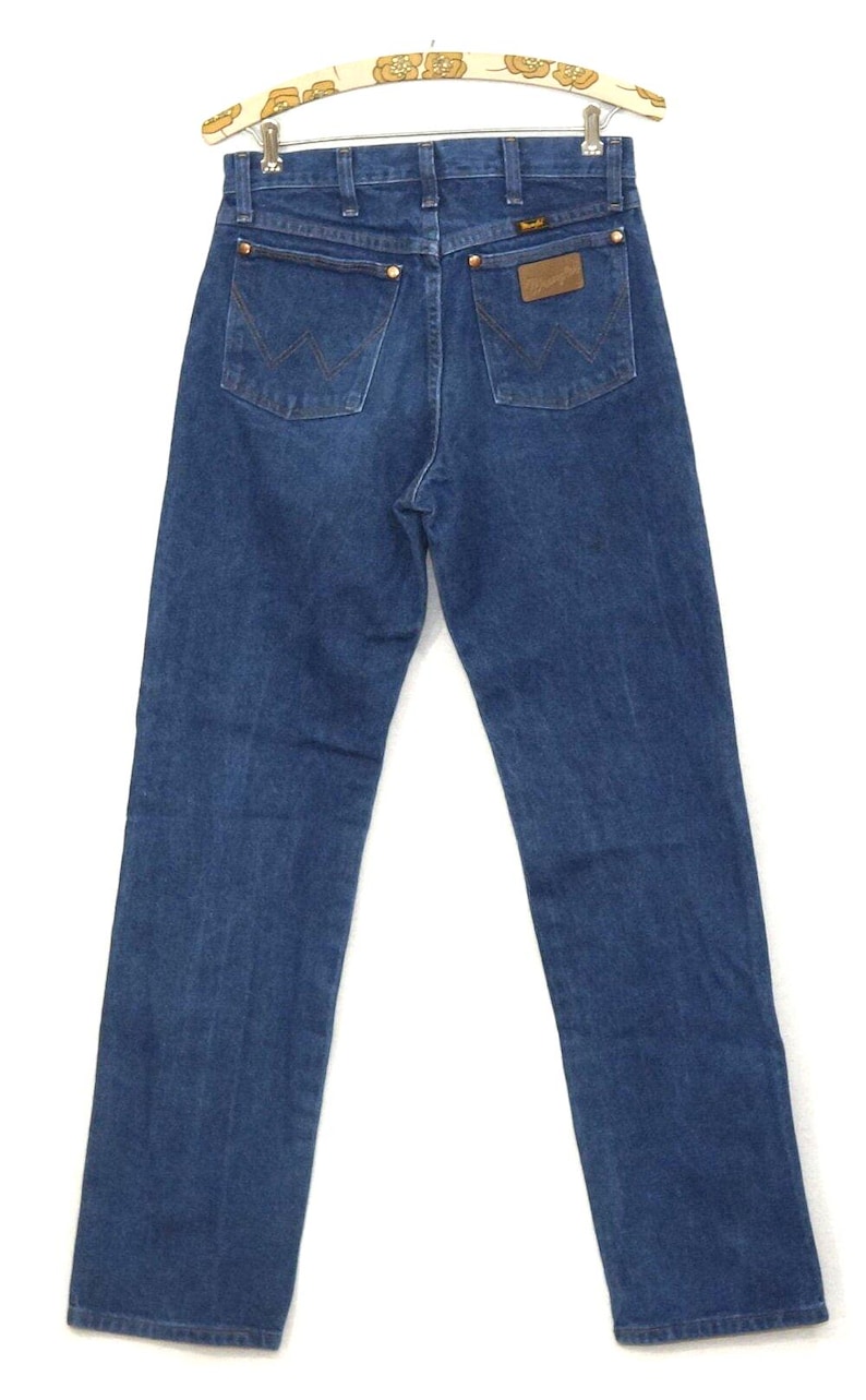 May include: Back view of blue denim jeans with the Wrangler logo on the back pocket and a brown leather patch. The jeans have classic five-pocket styling and are hanging on a vintage patterned hanger.