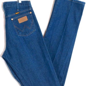 May include: Blue denim jeans with a classic straight leg cut. The jeans feature a brown leather patch on the back pocket with the Wrangler logo. The jeans are hung on a white hanger.