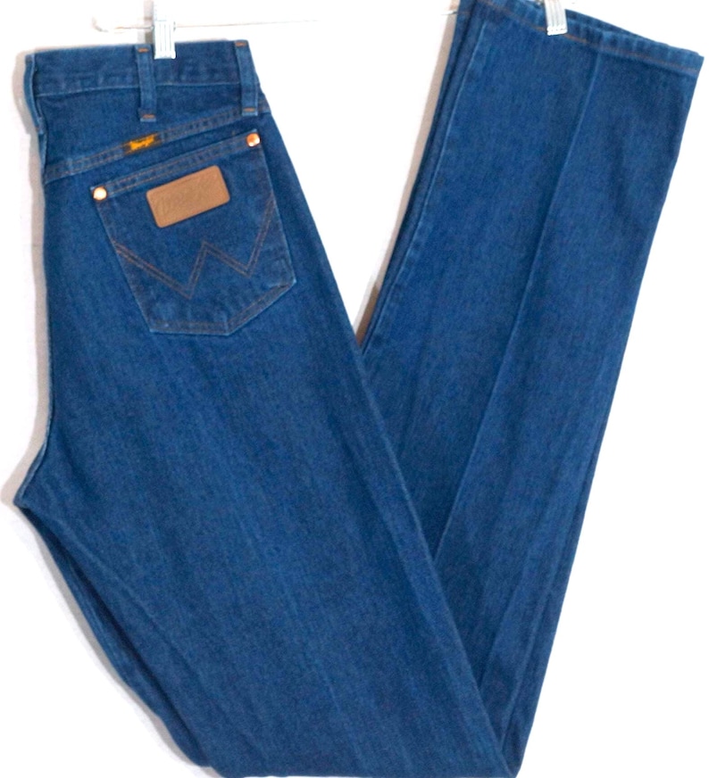May include: Blue denim jeans with a classic straight leg cut. The jeans feature a brown leather patch on the back pocket with the Wrangler logo. The jeans are hung on a white hanger.