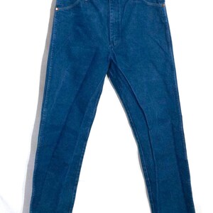 May include: A pair of blue denim jeans hanging against a white background. The jeans have a classic five-pocket design with copper-colored rivets and a button closure. The denim appears to be a medium weight.