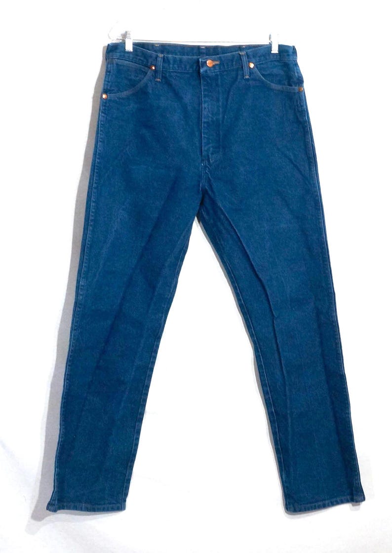 May include: A pair of blue denim jeans hanging against a white background. The jeans have a classic five-pocket design with copper-colored rivets and a button closure. The denim appears to be a medium weight.