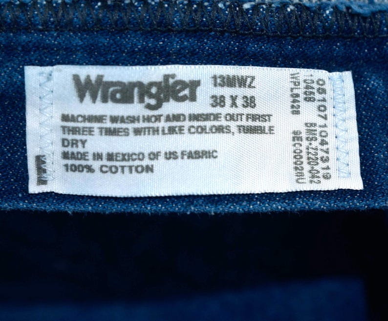 May include: Close-up of a blue denim fabric with a white label. The label reads "Wrangler" and includes washing instructions, size 38 x 38, and fabric details. The fabric is 100% cotton and made in Mexico.