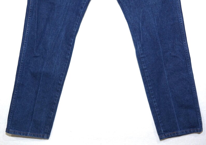 May include: Dark blue denim jeans, featuring a classic five-pocket design and a straight leg cut. The denim has a slightly faded appearance, with subtle wear marks. The jeans are laid flat on a white surface.
