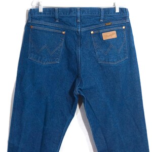 May include: Blue denim jeans with the Wrangler logo on the back pocket. The jeans have a classic five-pocket design with copper rivets and a leather patch. The jeans are hung on a white hanger.