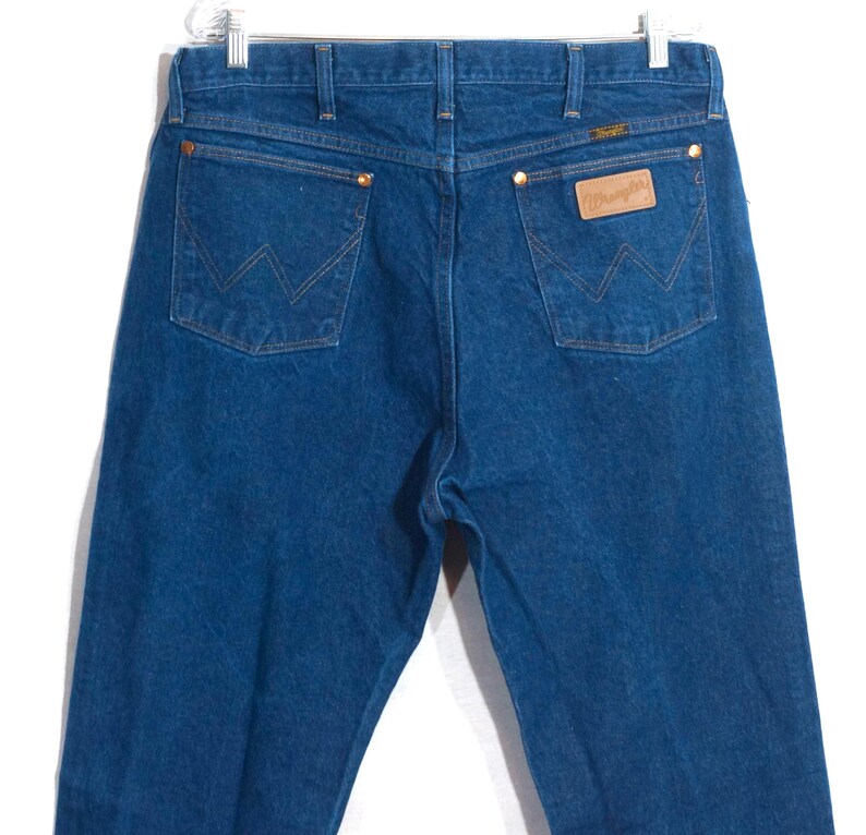May include: Blue denim jeans with the Wrangler logo on the back pocket. The jeans have a classic five-pocket design with copper rivets and a leather patch. The jeans are hung on a white hanger.