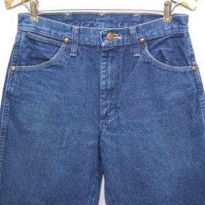 May include: Close-up of blue denim jeans with a classic five-pocket design. The jeans have a button and zipper closure, with copper-colored rivets and a brown leather patch. The jeans are hanging on a metal hanger.