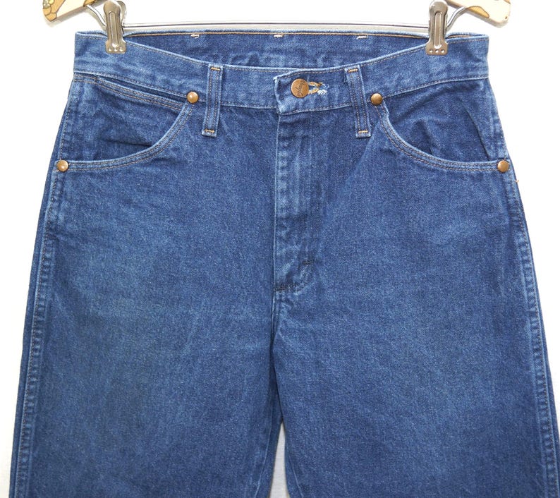 May include: Close-up of blue denim jeans with a classic five-pocket design. The jeans have a button and zipper closure, with copper-colored rivets and a brown leather patch. The jeans are hanging on a metal hanger.