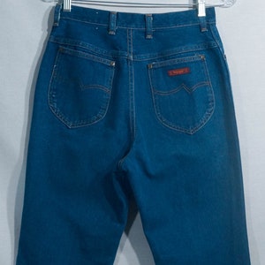 May include: Dark blue denim jeans with a high waist and classic five-pocket design. The back pockets have a unique curved shape, and a red "Wrangler" label is visible. The jeans are hanging on a clear hanger.