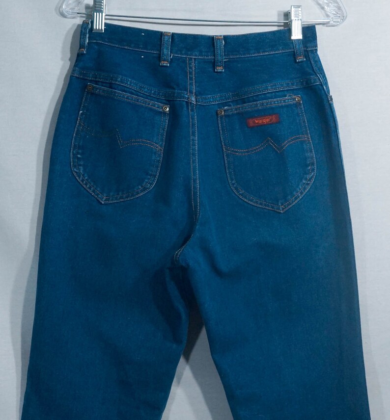 May include: Dark blue denim jeans with a high waist and classic five-pocket design. The back pockets have a unique curved shape, and a red "Wrangler" label is visible. The jeans are hanging on a clear hanger.