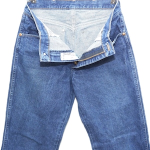 May include: Blue denim jeans with a partially unzipped fly and a button closure. The jeans are hanging on a metal hanger. The Wrangler brand label is visible.