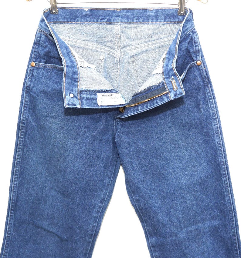 May include: Blue denim jeans with a partially unzipped fly and a button closure. The jeans are hanging on a metal hanger. The Wrangler brand label is visible.