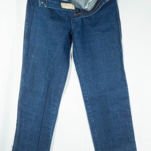 May include: Dark blue denim jeans, displayed open and hanging. The jeans have a classic five-pocket design, a button and zipper closure, and a small label. The denim appears to be a medium weight.