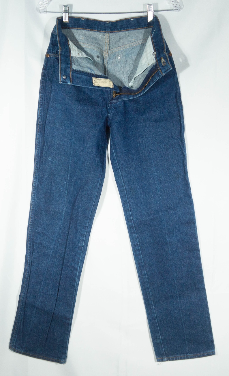 May include: Dark blue denim jeans, displayed open and hanging. The jeans have a classic five-pocket design, a button and zipper closure, and a small label. The denim appears to be a medium weight.
