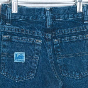 May include: A pair of dark blue denim jeans featuring a classic five-pocket design, a waistband with belt loops, and a light blue rectangular patch on the back pocket with the text Lee Authentic printed in a darker blue font.