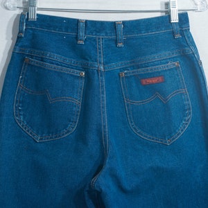 May include: Dark blue denim jeans with a high waist and Wrangler label. The jeans have classic five-pocket styling with copper-colored stitching and a brown leather patch. The back pockets feature a distinctive curved design.