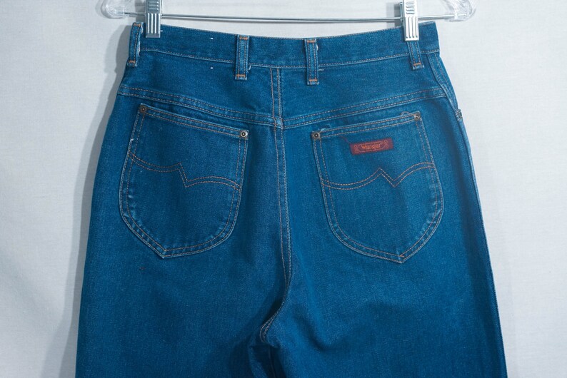 May include: Dark blue denim jeans with a high waist and Wrangler label. The jeans have classic five-pocket styling with copper-colored stitching and a brown leather patch. The back pockets feature a distinctive curved design.