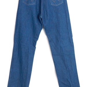 May include: Blue denim jeans with the Wrangler logo on the back pocket and a leather patch. The jeans have a classic five-pocket design and a straight leg fit. The jeans are hanging on a wooden hanger.