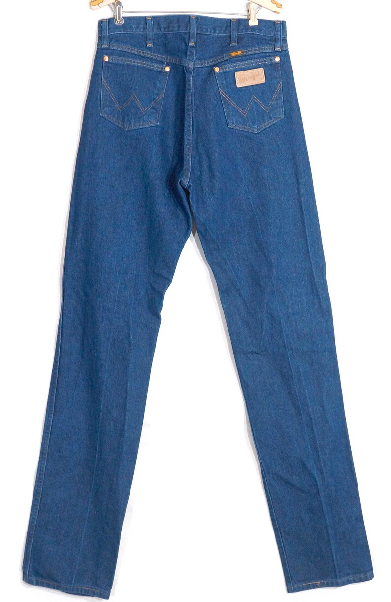 May include: Blue denim jeans with the Wrangler logo on the back pocket and a leather patch. The jeans have a classic five-pocket design and a straight leg fit. The jeans are hanging on a wooden hanger.