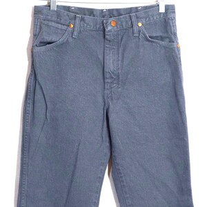 May include: A pair of dark gray denim jeans hanging from a metal hanger. The jeans have a classic five-pocket design with copper-colored rivets and button closures. The fabric appears to be a medium-weight denim.