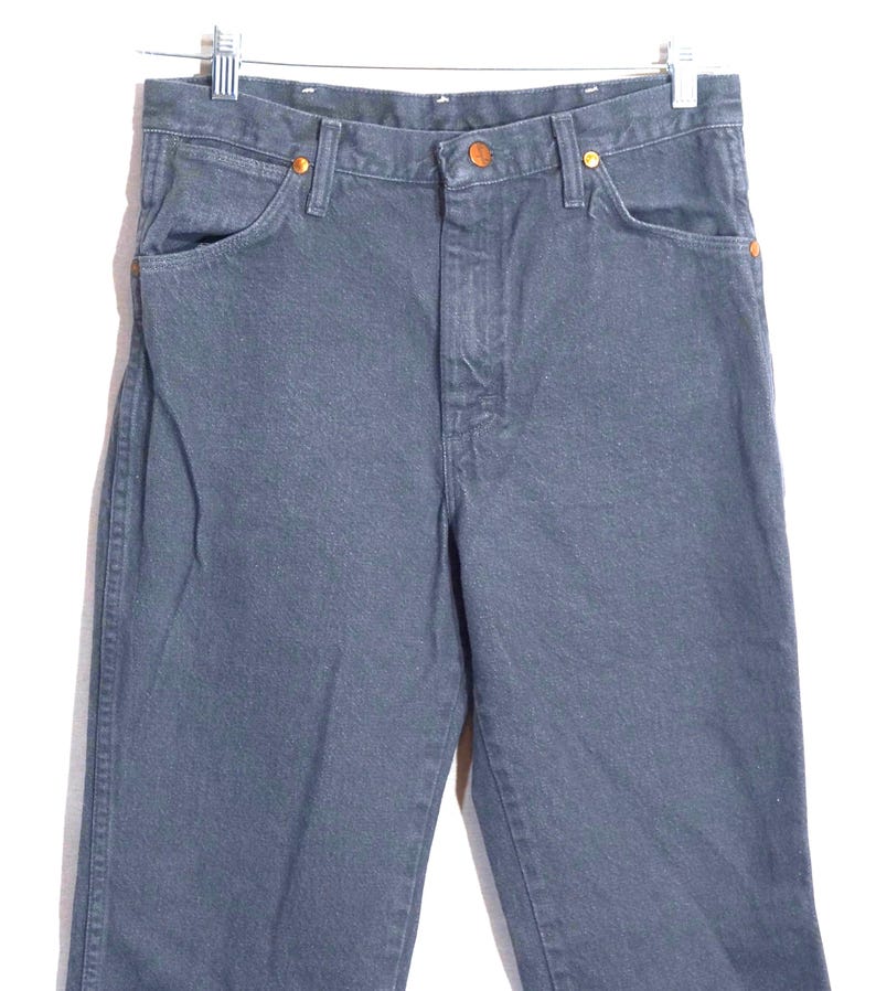 May include: A pair of dark gray denim jeans hanging from a metal hanger. The jeans have a classic five-pocket design with copper-colored rivets and button closures. The fabric appears to be a medium-weight denim.