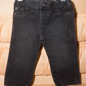 May include: Black denim pants for a child, featuring a button closure, belt loops, and classic five-pocket styling. The pants are hanging on a hanger, showcasing the details of the dark wash denim and stitching.