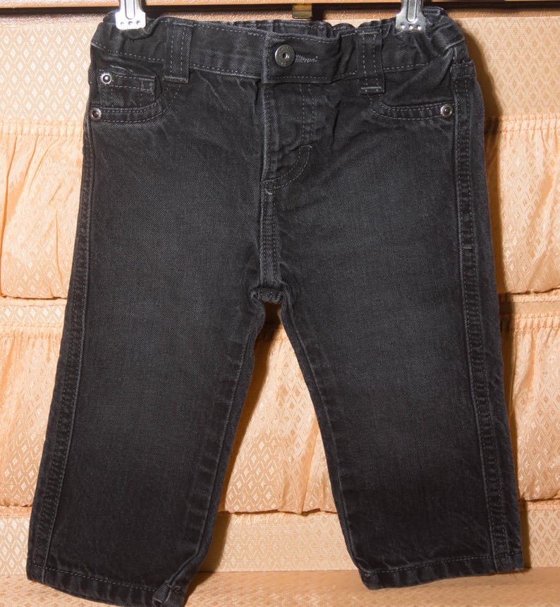 May include: Black denim pants for a child, featuring a button closure, belt loops, and classic five-pocket styling. The pants are hanging on a hanger, showcasing the details of the dark wash denim and stitching.