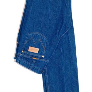 May include: Blue denim jeans hanging on a wooden hanger. The jeans have a classic five-pocket design, a brown leather patch, and a distinctive back pocket stitch. The denim is a medium wash, and the jeans appear to be unworn.