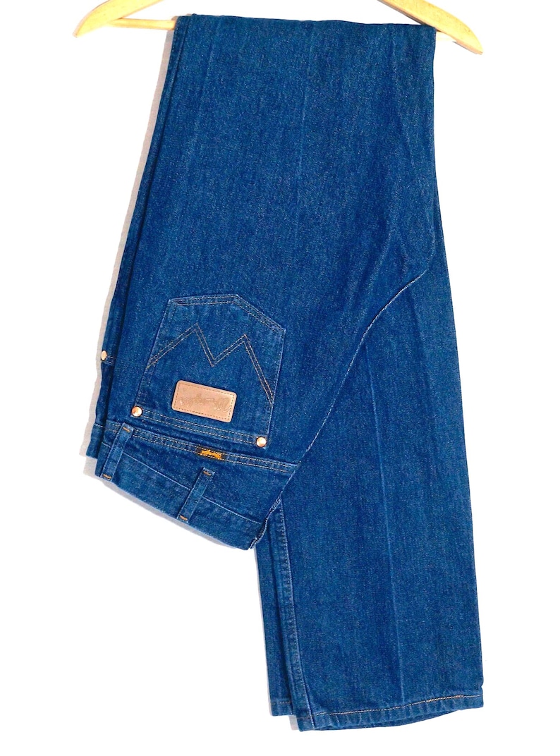 May include: Blue denim jeans hanging on a wooden hanger. The jeans have a classic five-pocket design, a brown leather patch, and a distinctive back pocket stitch. The denim is a medium wash, and the jeans appear to be unworn.