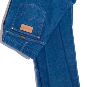 May include: A pair of blue denim jeans hanging on a wooden hanger. The jeans have a classic five-pocket design, with a brown leather patch on the back pocket. The back pocket features a stitched design. The jeans are a medium wash.