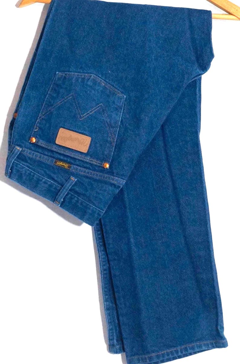 May include: A pair of blue denim jeans hanging on a wooden hanger. The jeans have a classic five-pocket design, with a brown leather patch on the back pocket. The back pocket features a stitched design. The jeans are a medium wash.