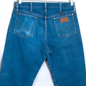 Puede incluir: Vaqueros azules de mezclilla con el logo de Wrangler en el bolsillo trasero. Los vaqueros tienen un dise&ntilde;o cl&aacute;sico de cinco bolsillos con remaches de cobre y un parche de cuero. Los vaqueros est&aacute;n colgados de una percha de metal.