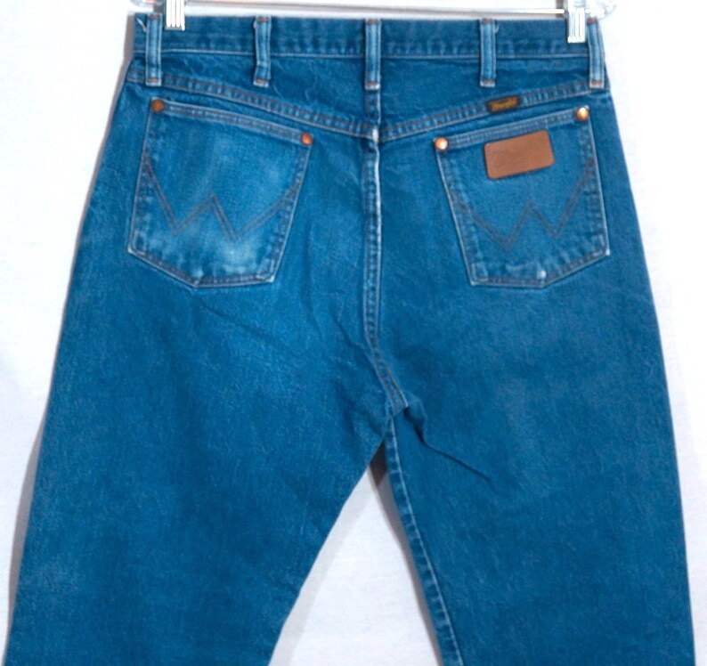 Puede incluir: Vaqueros azules de mezclilla con el logo de Wrangler en el bolsillo trasero. Los vaqueros tienen un dise&ntilde;o cl&aacute;sico de cinco bolsillos con remaches de cobre y un parche de cuero. Los vaqueros est&aacute;n colgados de una percha de metal.