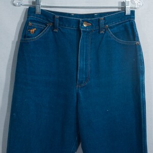 May include: Dark blue denim jeans with a classic five-pocket design. Features a small embroidered horse logo on the front pocket. The jeans are hanging on a metal hanger.