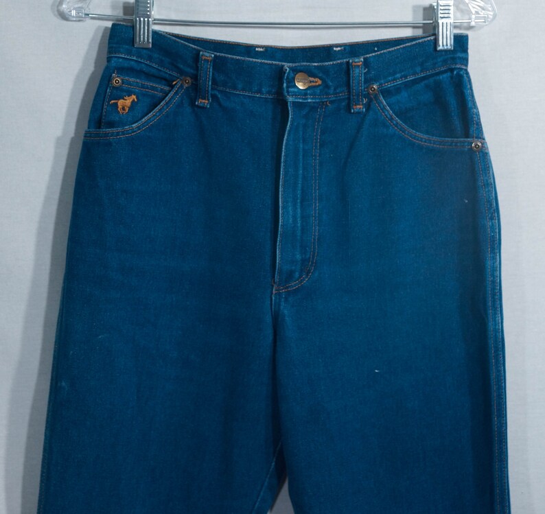 May include: Dark blue denim jeans with a classic five-pocket design. Features a small embroidered horse logo on the front pocket. The jeans are hanging on a metal hanger.