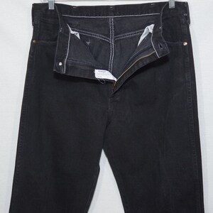 May include: Black denim jeans with a zipper fly and white stitching. The jeans are hanging on a metal hanger. The Wrangler brand label is visible.