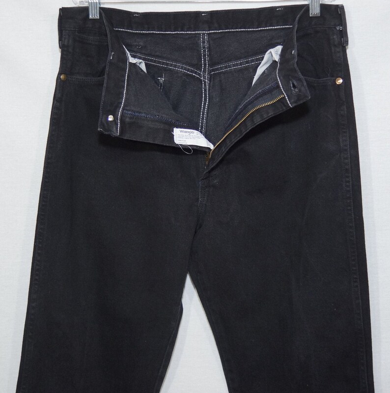 May include: Black denim jeans with a zipper fly and white stitching. The jeans are hanging on a metal hanger. The Wrangler brand label is visible.