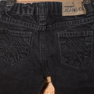 May include: Close-up of the back of black denim jeans. The jeans have two back pockets with a stitched 'W' design. A brown leather patch with the 'Wrangler JEANS CO' logo is visible. The jeans are hanging on a hanger.