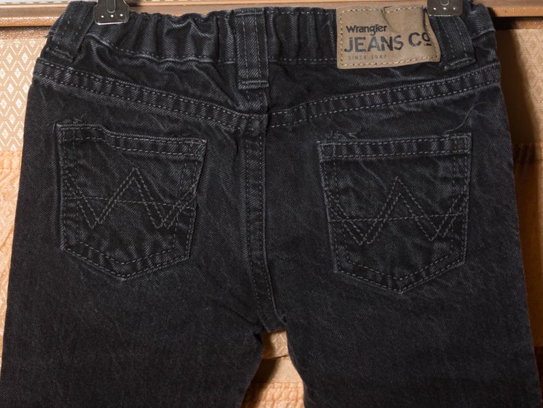 May include: Close-up of the back of black denim jeans. The jeans have two back pockets with a stitched 'W' design. A brown leather patch with the 'Wrangler JEANS CO' logo is visible. The jeans are hanging on a hanger.